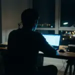 Silhouette of a person using a laptop in a low-light environment representing online anonymity.