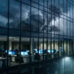 A modern glass office building reflecting a stormy sky with empty desks inside.